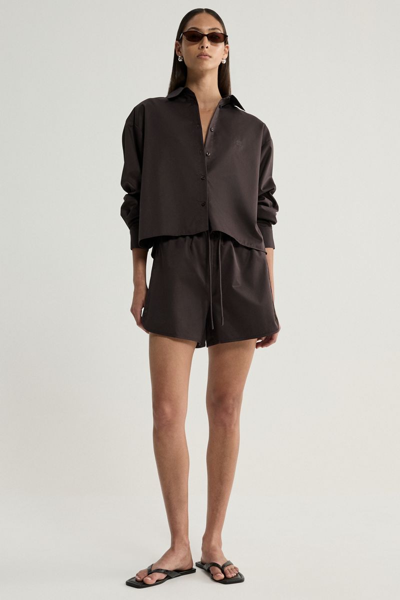 A model wearing a dark brown matching two-piece set consisting of a long-sleeve button-up shirt and shorts, styled with black sunglasses and black flat sandals against a light gray studio background.