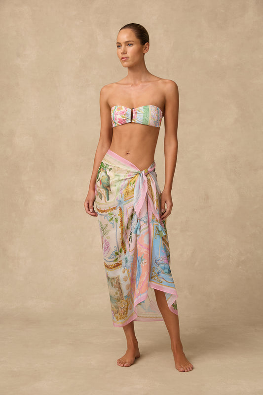 A woman stands barefoot against a warm beige textured backdrop, wearing a matching strapless bandeau top and sarong set with a pastel multicolor print featuring pink, blue, green, and cream tones.