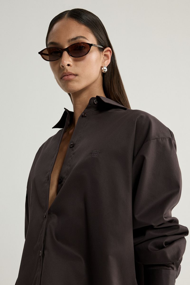 A woman wearing an oversized dark brown button-up shirt and dark sunglasses, photographed against a light gray studio background.