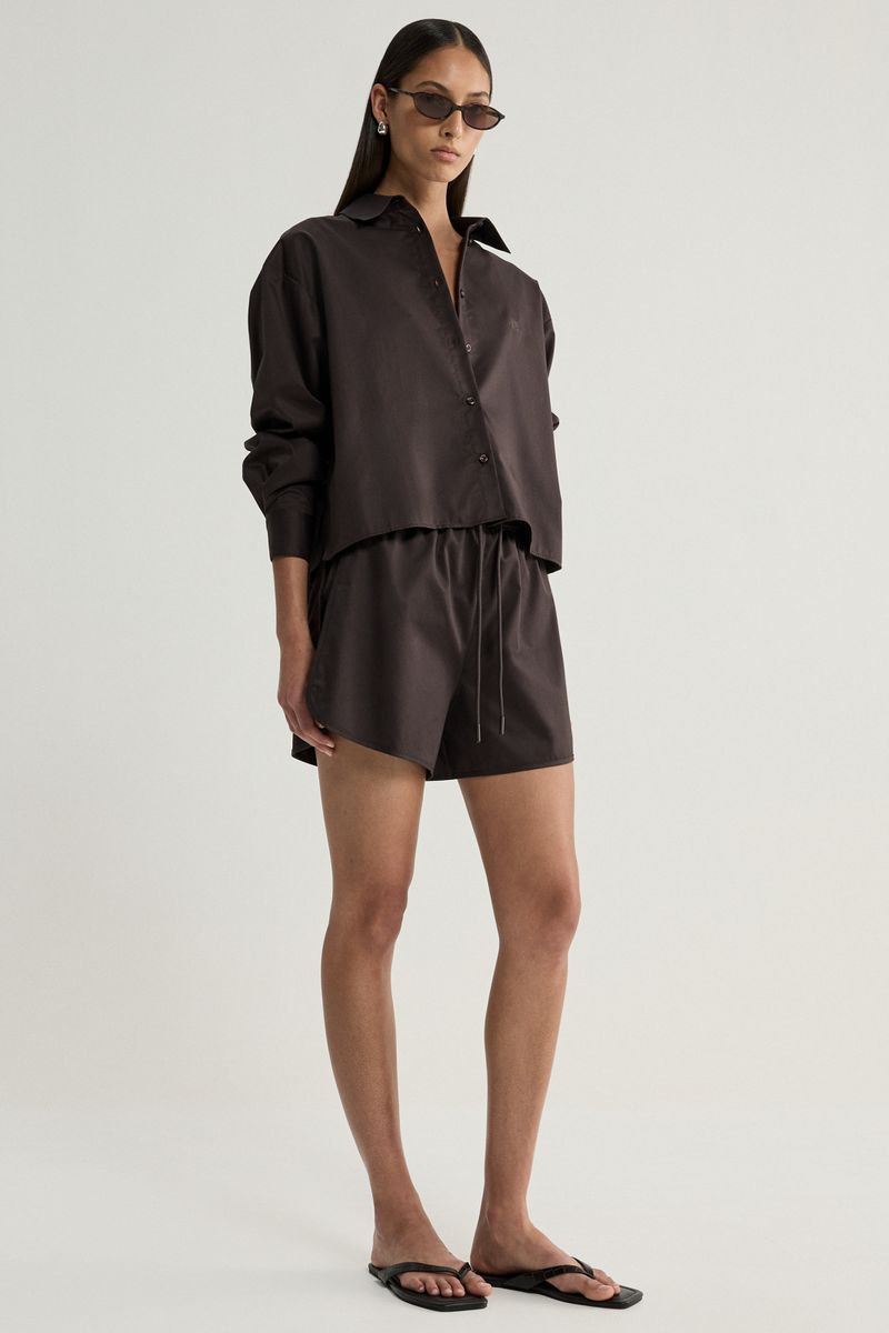 A model wearing a dark brown satin-like two-piece set consisting of a long-sleeve button-up shirt and matching shorts, styled with black sunglasses and black flip-flop sandals against a light gray studio background.