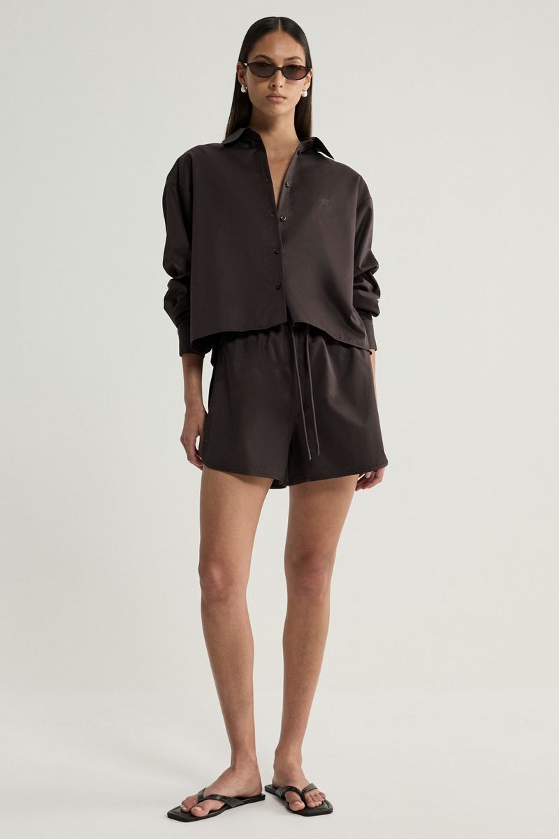 A model stands against a light gray studio background wearing a dark brown satin-like two-piece set consisting of a long-sleeve button-front shirt and matching shorts with a drawstring, along with black sandals and sunglasses.