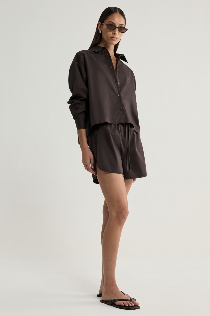 A model wearing a dark brown matching lounge set consisting of a long-sleeve button-down shirt and shorts, styled with sunglasses and black flip-flop sandals against a plain light gray studio background.