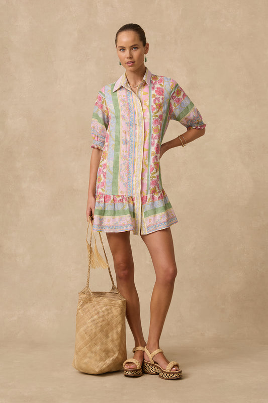 A woman models a short-sleeve button-up shirt dress with a pastel multicolor floral and stripe pattern, paired with tan platform sandals and holding a woven straw tote bag against a textured beige studio backdrop.