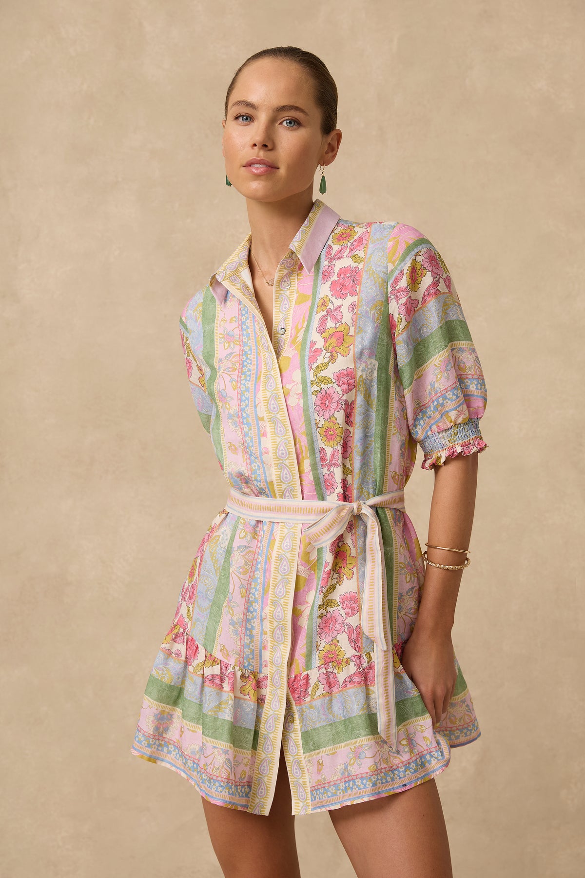 A woman modeling a short, pastel multicolor patterned wrap dress or robe-style shirt dress with puff sleeves and a tie waist, photographed against a warm beige studio backdrop.