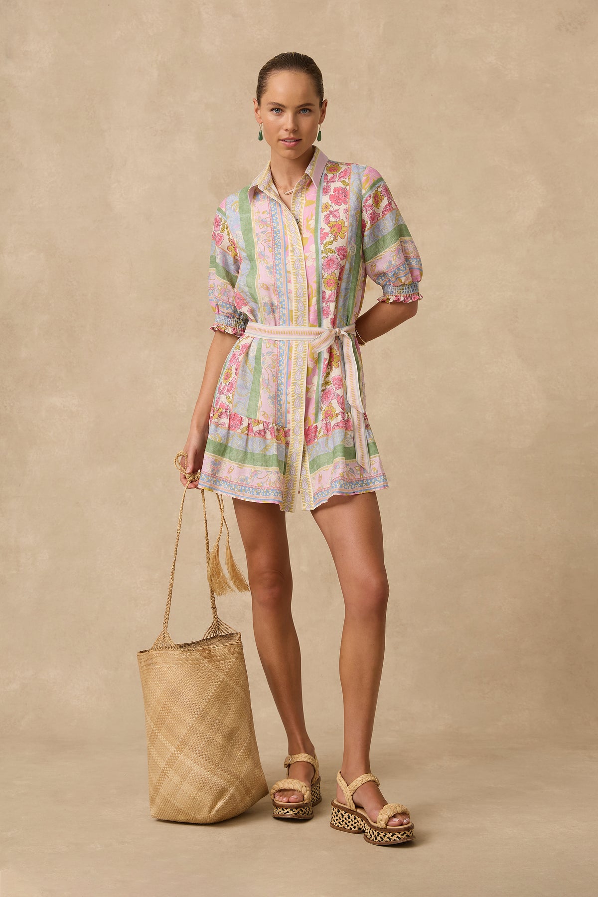 A woman stands against a warm beige textured backdrop wearing a short, colorful floral-and-striped print shirt dress with puff sleeves and a tie waist. She is holding a large woven straw tote bag and wearing beige platform sandals.