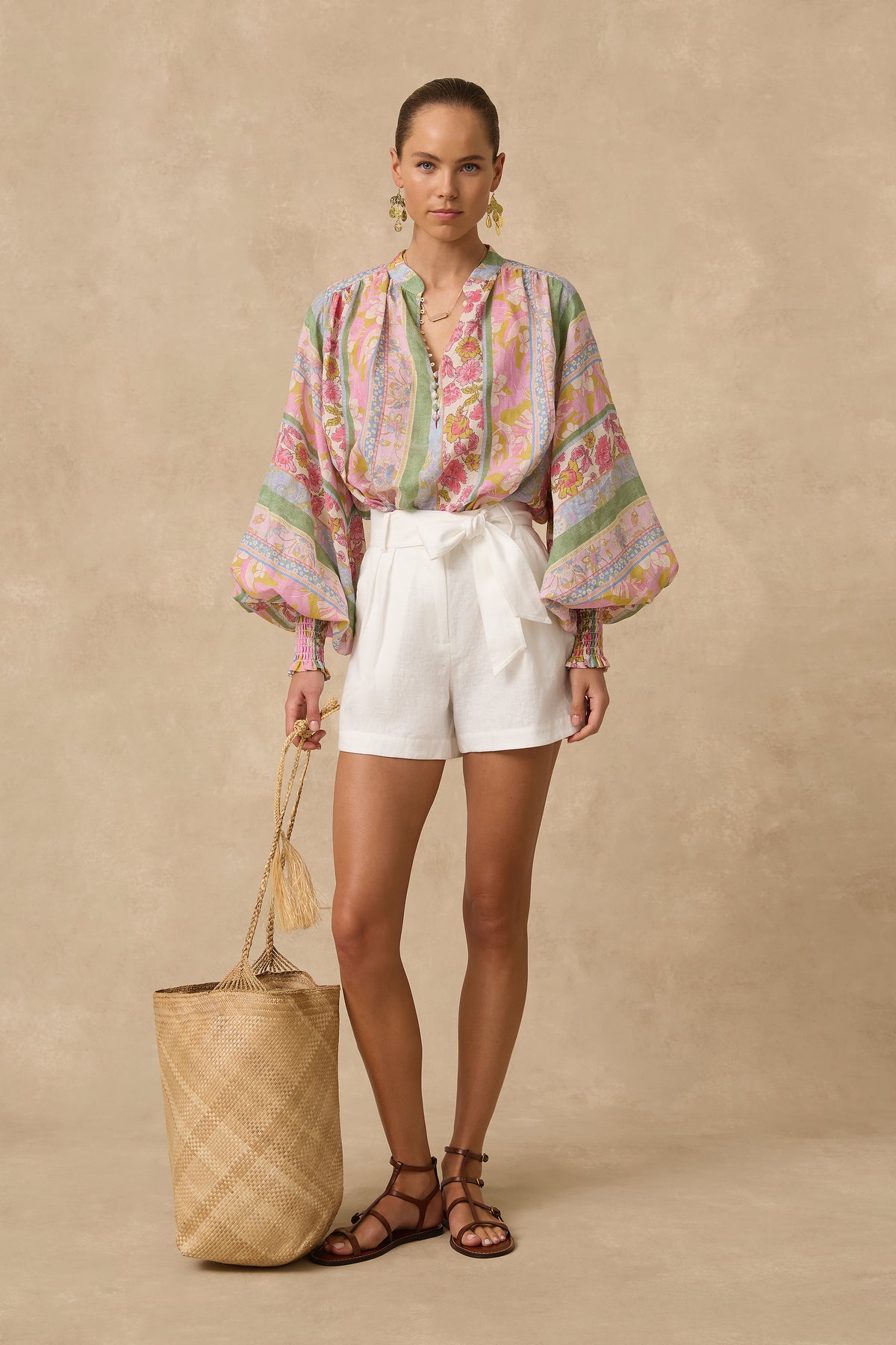 A woman stands against a warm beige studio backdrop wearing a colorful paisley blouse with voluminous sleeves, white shorts, brown strappy sandals, and holding a large woven straw tote bag.