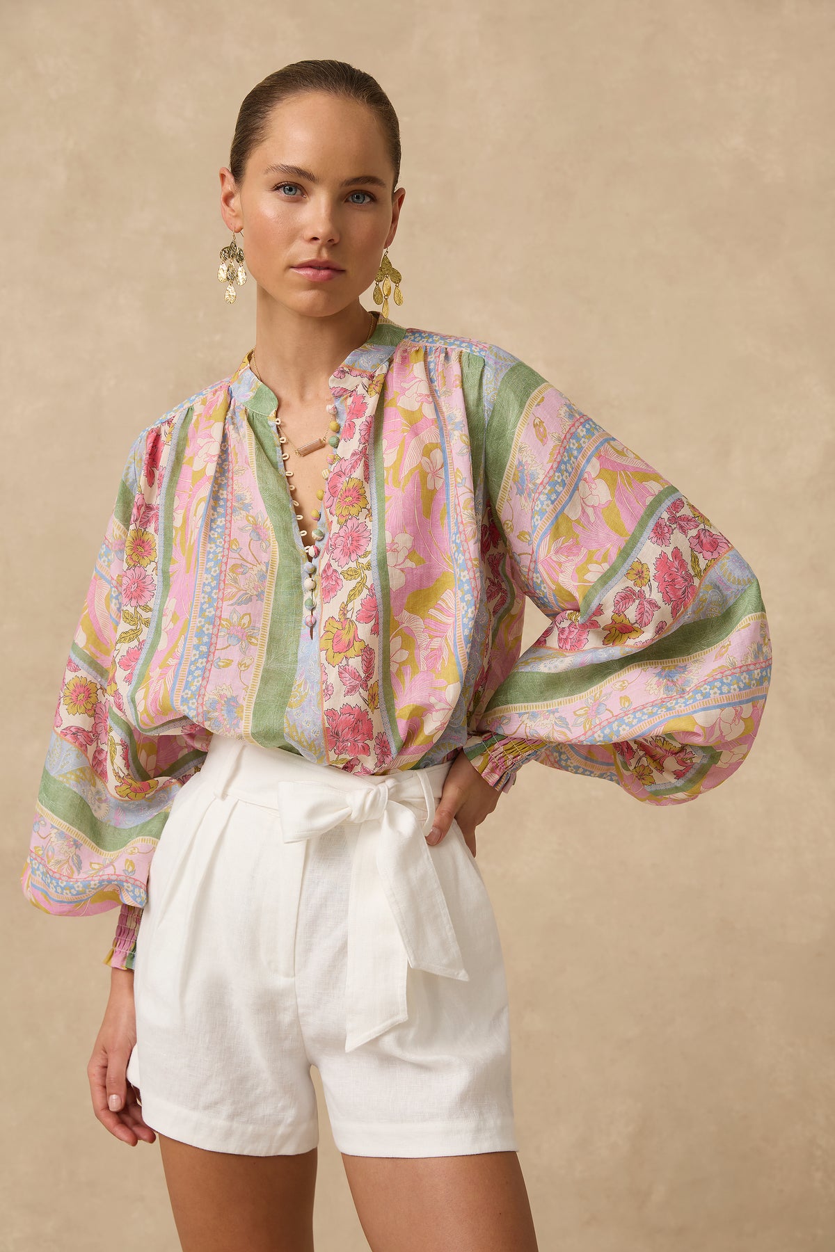 A woman modeling a long-sleeve blouse with a colorful paisley and floral print in pink, green, blue, and yellow, paired with white shorts, against a warm beige studio background.