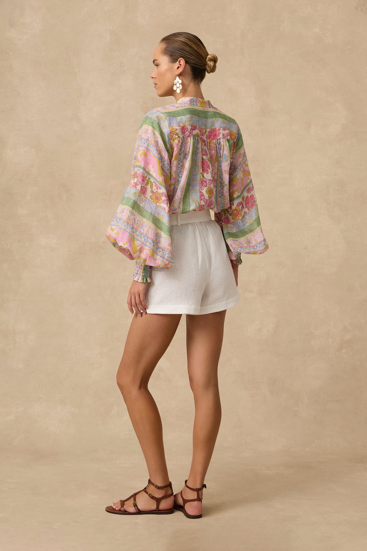 A woman is shown from the back wearing a colorful floral long-sleeve blouse with green stripes and white shorts, standing against a textured beige studio backdrop.