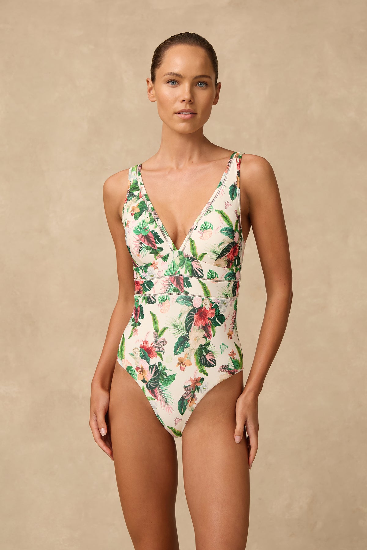 A woman modeling a one-piece tropical print swimsuit with a deep V-neckline, set against a textured beige studio backdrop.