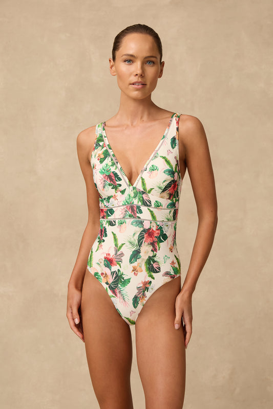 A woman modeling a one-piece tropical print swimsuit with a deep V-neckline, set against a textured beige studio backdrop.