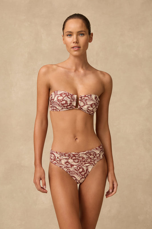 A woman modeling a two-piece swimsuit with a red and cream paisley pattern against a textured beige studio 