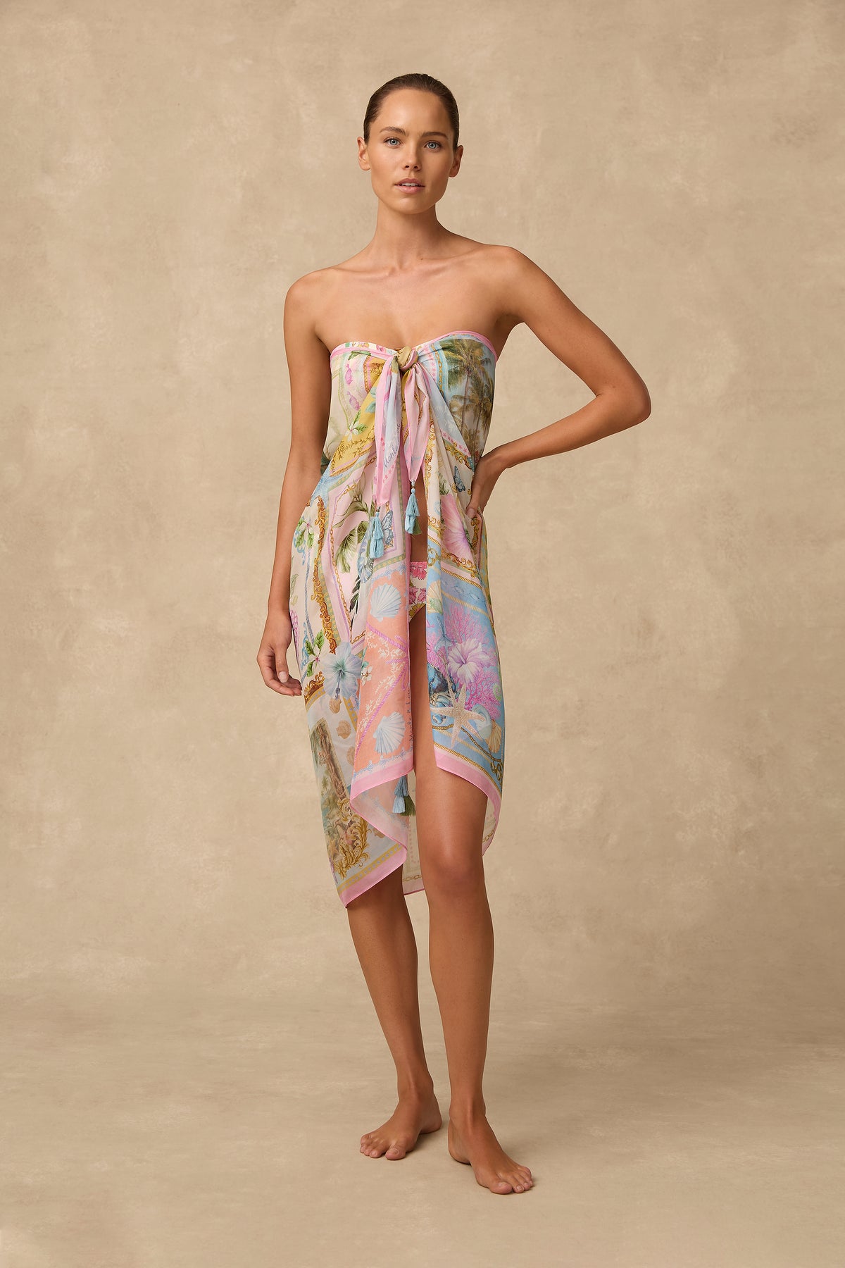 A woman stands barefoot against a warm beige textured backdrop wearing a strapless tie-front sarong dress with a pastel multicolor tropical print.
