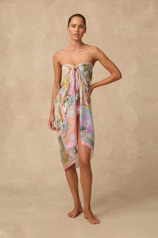 A woman stands barefoot against a warm beige textured backdrop wearing a strapless tie-front sarong dress with a pastel multicolor tropical print.