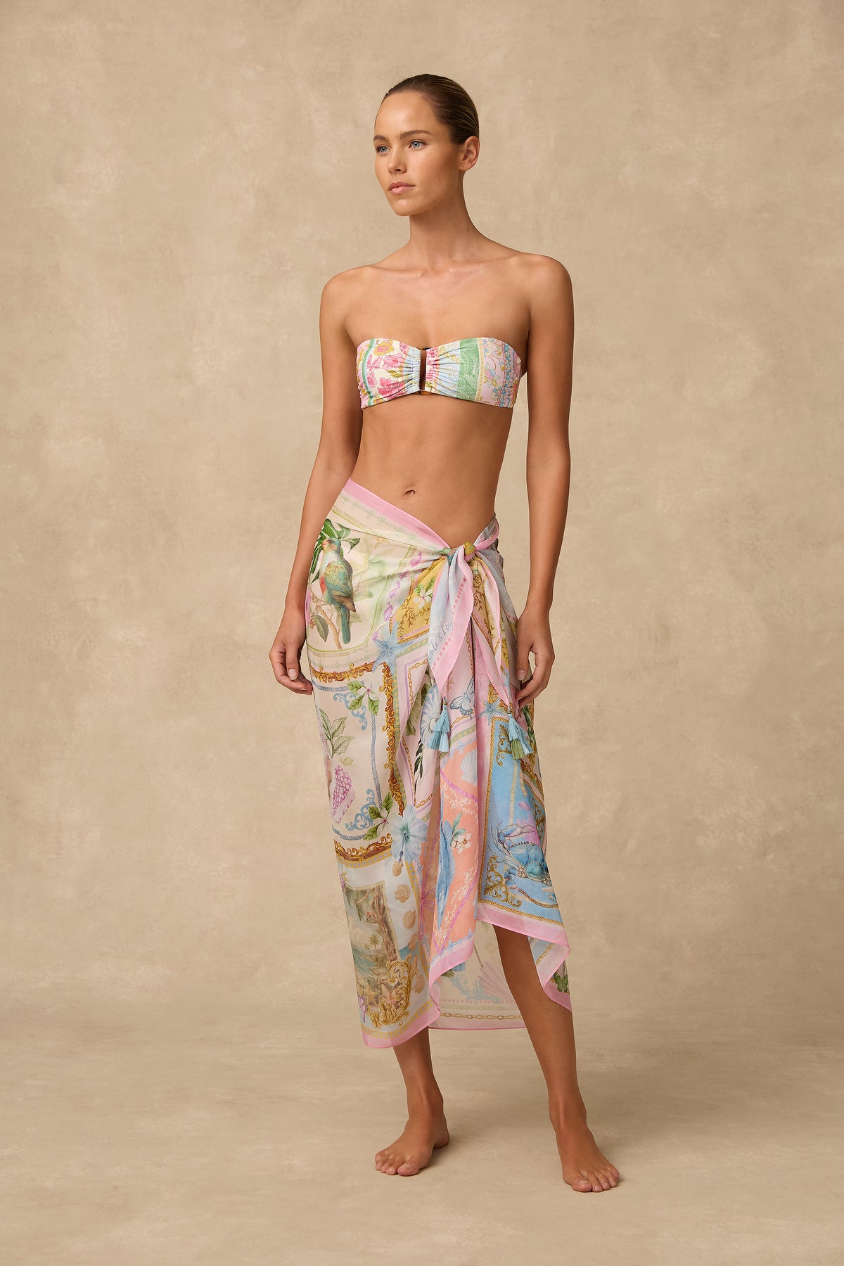 A woman stands barefoot against a warm beige textured backdrop, wearing a matching strapless bandeau top and sarong set with a pastel multicolor print featuring pink, blue, green, and cream tones.